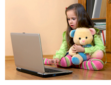 Photo of girl holding her stuffed animal while playing on a laptop computer